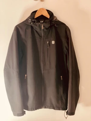 Svart vindjacka från 8848 Altitude - Svart vindjacka från 8848 Altitude med huva, dragkedja framtill och praktiska fickor med dragkedja. Snyggt broderad 8 på bröstet och justerbar nederkant med snörning. Perfekt för dig som gillar stilren och funktionell design.