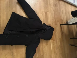 Svart dunjacka från Canada Goose - Säljer en svart dunjacka från Canada Goose med huva och klassisk logga på ärmen. Jackan har dragkedja och stora knappar framtill samt en ficka med dragkedja på ärmen. Perfekt för kalla dagar och riktigt snygg streetstil.