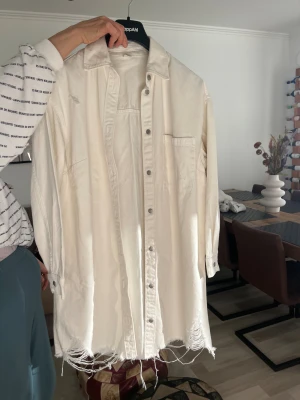 Vit overshirt från &Denim, S - Vit overshirt från &Denim i storlek S. Skjortan har klassisk krage, knappstängning och en bröstficka. Materialet är bomull och nederkanten har en snygg fransad finish som ger en cool vibe. Perfekt att styla lager på lager för en avslappnad look.