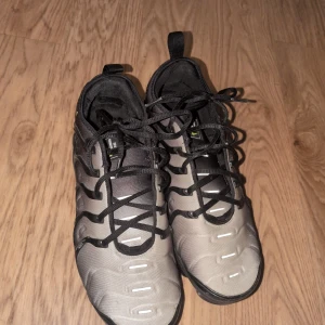 Nike Air Vapormax sneakers grå/svart - Nike Air Vapormax sneakers i storlek 43 med grå och svart ovandel, svarta snören och coola detaljer. Sulan är transparent svart med synliga luftkuddar och gröna Nike-loggor. Ovandelen är i syntetmaterial med mönstrade linjer och reflekterande inslag. Perfekt för dig som gillar sportig och modern stil.