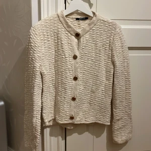Beige cardigan jacka från Gina Tricot - Superfin beige cardigan jacka från Gina Tricot med strukturerad yta och snygga guldiga knappar framtill. Jackan har rundad halsringning och långa ärmar, perfekt för lager-på-lager och en clean look. Knappt använd.