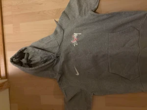 Grå Stüssy x Nike hoodie med tryck - Säljer en grå hoodie från Stüssy x Nike med stort färgglatt Stüssy-tryck på ryggen och mindre tryck på bröstet. Tröjan har huva med snörning, magficka och långa ärmar. Perfekt för dig som gillar streetwear och vill sticka ut med coola detaljer.