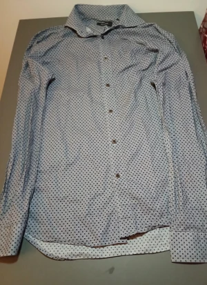 Blå prickig skjorta med knappar - Snygg blå skjorta med små svarta prickar och klassisk krage. Skjortan har lång ärm och knäppning framtill med svarta knappar. Perfekt för dig som gillar stilrena mönster och vill ha en skjorta som sticker ut lite. Materialet känns lätt och bekvämt mot huden.
