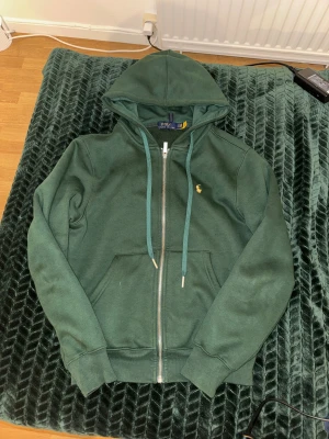 Grön hoodie från Polo Ralph Lauren - Mörkgrön hoodie från Polo Ralph Lauren med gul broderad logga på bröstet. Tröjan har huva med snörning, dragkedja framtill och två fickor. Insidan är mjuk och fleecefodrad, perfekt för kyliga dagar. Klassisk och stilren design med ribbade muddar.