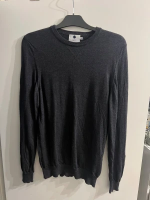 Mörkgrå stickad tröja från NN07 Medium - Snygg mörkgrå stickad tröja från NN07 i 100% extrafin ull. Rund halsringning med ribbade muddar vid ärmslut och nederkant. Perfekt för dig som gillar stilrena och enkla plagg med skön passform.
