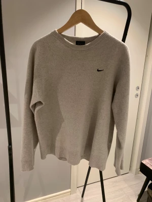 Beige stickad tröja från Nike vintige  - Snygg beige stickad tröja från Nike med diskret svart logga på bröstet. Tröjan har rund hals, ribbade muddar och lång ärm. Perfekt för dig som gillar enkel och clean stil med sportig touch.