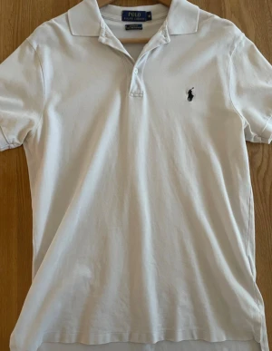 Vit pikétröja från Polo Ralph Lauren - Vit pikétröja från Polo Ralph Lauren i klassisk modell med krage och två knappar. Tröjan har det ikoniska broderade logot på bröstet i svart. Tillverkad i mjuk och stretchig pima bomull för extra komfort. Perfekt för en clean och stilren look.