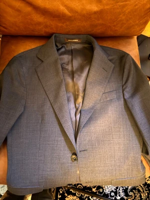 Grå kavaj från Oscar Jacobson - Snygg grå kavaj från Oscar Jacobson i klassisk modell med ett knappslå och diskret struktur. Kavajen har smal krage, innerfoder och flera fickor fram. Perfekt för dig som vill ha en stilren och modern look.