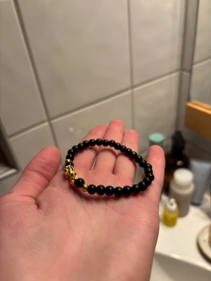 Svart Pärlarmband - Snyggt armband med svarta runda pärlor och detaljer i guldfärg, inklusive en cool drake. Perfekt för dig som gillar unika accessoarer med lite edge. Elastiskt band gör det enkelt att ta på och av.
