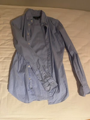 Blå slim fit skjorta från Ralph Lauren - Snygg ljusblå skjorta från Ralph Lauren i slim fit-modell. Klassisk design med knappar framtill och broderad logga på bröstet. Skjortan har lång ärm och är gjord i mjuk bomull som känns skön mot huden. Perfekt för dig som gillar stilrena och tidlösa plagg.