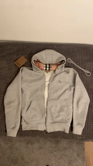Grå Burberry hoodie med dragkedja - Snygg grå hoodie från Burberry med klassiskt rutigt foder i huvan. Tröjan har dragkedja framtill, två fickor och ribbade muddar. Ikonisk Burberry-logga broderad på bröstet. Perfekt för dig som vill ha en stilren och lyxig streetwear-look.