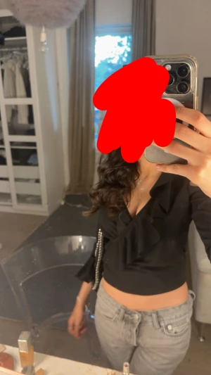 Svart omlottopp med volang  - Svart omlottopp  med volangdetaljer längs v-ringningen och axelpartiet. Toppen har långa ärmar med öppna axlar och lätt utsvängda ärmslut. Croppad passform som ger en snygg siluett. Perfekt att styla med jeans eller kjol.