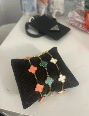 Färgglatt fyrklöverarmband i guld - Snyggt armband med fyrklöverformade berlocker i guld. Berlockerna är i färgerna korallrosa, grön och vit, och har en elegant guldkant runt varje klöver. Kedjan är tunn och guldfärgad, vilket ger ett lyxigt och trendigt intryck. Perfekt för att addera färg till din outfit.