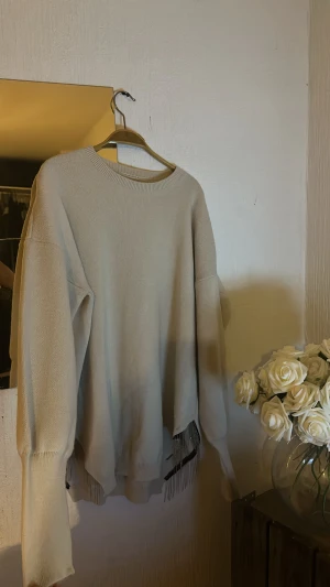 Beige stickad tröja med sliverdetaljer på sidorna - Beige stickad tröja med oversized passform och långa ärmar. Tröjan har rund halsringning och coola fransdetaljer i metall längs nederkanten som ger en unik touch. Perfekt för dig som gillar en chill och trendig stil.