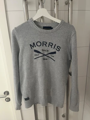 Grå stickad tröja från Morris - Snygg grå stickad tröja från Morris med mörkblå tryck och broderad text på bröstet. Rund halsringning och ribbade muddar vid ärmslut och nederkant. Perfekt för dig som gillar klassisk stil med en sportig touch.