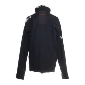 Mörkblå vindjacka från Helly Hansen - Mörkblå vindjacka från Helly Hansen med vit dragkedja och röda detaljer på fickorna. Jackan har hög krage med HH-logga, tre framfickor med dragkedja och justerbar nederkant. Materialet är polyester och polyuretan, vilket gör den lätt och funktionell.