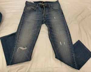 Lee Daren jeans W31 L34 blå - Snygga Lee Daren jeans i klassisk blå tvätt med slitningar och lätt distressed look. Modellen har raka ben, fem fickor och knappgylf. Jeansen är tillverkade i bomull och har en normal passform som funkar till de flesta stilar.
