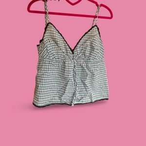 Rutig linnetopp med spets och knyt - Supersöt linnetopp från Cotton on med bruna, vita och blå rutor, tunna axelband och v-ringad hals. Svart spetskant längs halsringningen och nederkant ger extra detalj. Knyt framtill och lätt rynkad midja. 