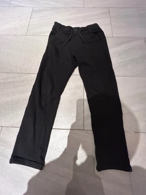 Svarta kostymbyxor med snöre - Svarta kostymbyxor med rak passform och snörning i midjan. Byxorna har elastisk midja, knapp framtill och två bakfickor. Materialet är mjukt och bekvämt, perfekt för chill dagar. Snygg och enkel design som funkar till det mesta. Mjukt material!