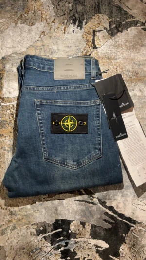 Stone Island jeans - Snygga blå jeans från Stone Island med klassisk passform och ikonisk kompasspatch på bakfickan. Jeansen har tydliga kontrastsömmar och är tillverkade i slitstarkt denim. Perfekt för dig som vill ha en cool och tidlös look med streetkänsla.