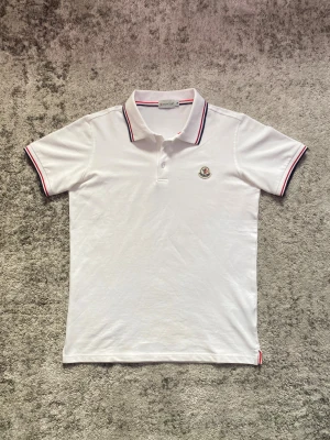 Vit Moncler Piké  - Riktigt stilren Pikétröja med blåa och röda ränder på ärmar och krage. Storlek M men sitter mer som en S med den tighta passformen. Hör gärna av dig vid minsta fundering!