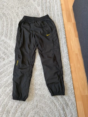 Nike NOCTA svarta träningsbyxor S - Säljer ett par svarta Nike NOCTA träningsbyxor i storlek S. Byxorna har gul NOCTA-logga på benet och gul swoosh på höften. Tillverkade i polyester med elastisk midja och muddar vid benslut. Perfekta för dig som gillar streetwear och sportig stil.