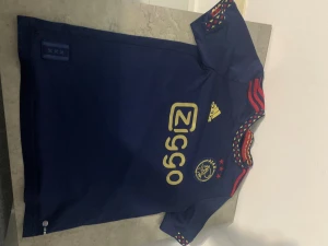 Ajax blå fotbollströja Adidas 13-14Y - Ajax fotbollströja i mörkblått från Adidas med guldfärgade tryck och logga. Röd-gula detaljer på kragen och röda ränder på axlarna. Materialet är lätt och ventilerande polyester, perfekt för träning eller match. Ziggo sponsortryck på bröstet och Ajax-emblem med tre stjärnor.