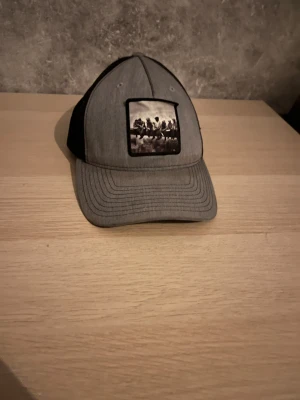 Grå keps från Richardson - Snygg grå keps från Richardson med svart mesh på baksidan och ett svartvitt tryck framtill. Kepsen har böjd skärm och justerbar passform. Perfekt för en avslappnad stil.