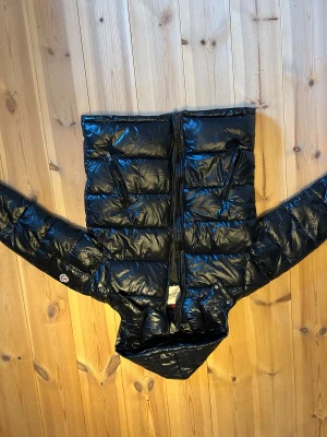 Svart dunjacka från Moncler - Säljer en svart, glansig dunjacka från Moncler med huva och två snedställda dragkedjefickor. Jackan har klassisk Moncler-logga på ärmen och coolt serietryck på insidan. Perfekt för dig som vill ha en varm och snygg jacka med street vibe. Priset inte hugget i sten.