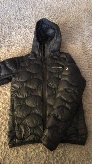 Svart dunjacka Peak Performance  - Svart dunjacka från Peak Performance i storlek S. Jackan har huva, snygg quiltad design och dragkedja framtill. Perfekt för kalla dagar och har en stilren look med diskret logga på bröstet och ärmen. Passar dig som gillar funktion och enkel stil.