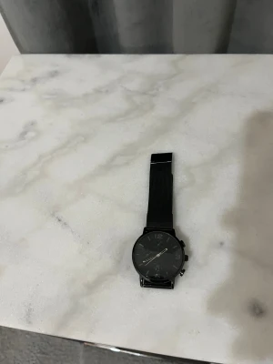 Svart stilren klocka med meshband - Snygg svart klocka med rund urtavla och svart mesharmband. Klockan har minimalistisk design med tydliga siffror och flera små visare på urtavlan. Metallspänne och mattsvart finish ger en modern vibe. Perfekt för dig som gillar stilrena accessoarer.