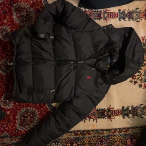 Svart pufferjacka från Polo Ralph Lauren - Svart pufferjacka från Polo Ralph Lauren med huva och dragkedja framtill. Jackan har en klassisk, quiltad design och en liten röd logga på bröstet. Perfekt för kalla dagar och har flera praktiska fickor med dragkedja.