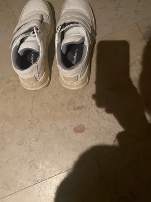 Vita sneakers från Acne Studios - Säljer ett par vita sneakers från Acne Studios med  platt sula och två breda kardborreband över foten. Skorna har rund tå med den klassiska smiley loggan baktill. Tillverkade i skinn med grå insida och beigeaktiga sulor. Jag har vanligtvis storlek 38 men dem passar mig bra trots att det är storlek 37. Obs! Det förekommer ytligt smuts, då dom inte är tvättade på bilden, men sådant som går bort enkelt, såklart är dem avtvättade innan jag skickar paketet. Använda, men mycket bra skick!