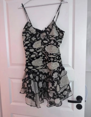 Svart blommig kortklänning med volang - Supersöt svart kortklänning med vita blommor och tunna axelband. Klänningen har volangig kjol och ett snyggt blommigt mönster som ger en romantisk vibe. Perfekt för dig som gillar en feminin och trendig stil.