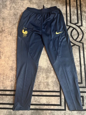Blå Frankrike Nike träningsbyxor FFF - Snygga marinblå träningsbyxor från Nike med FFF-logga och gul swoosh på framsidan. Byxorna har elastisk midja med snörning och smal passform. Materialet är polyester och har subtila linjer på benen för extra stilpoäng.