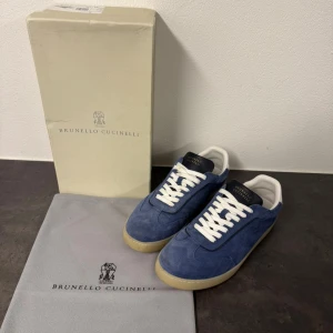 Blå sneakers Brunello Cucinelli - Snygga blå sneakers från Brunello Cucinelli i mocka med vita skinn-detaljer och vit snörning. Yttersulan är beige och har en platt design. Logotypen är tryckt i guld på hälen och plösen. Perfekt för dig som gillar stilrena och lyxiga sneakers. Storlek 43