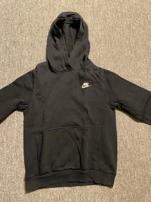 Svart Nike hoodie - Svart hoodie från Nike med klassisk vit logga på bröstet. Tröjan har huva, lång ärm och en stor magficka framtill. Tillverkad i mjuk bomullsmix, perfekt för chill eller streetwear. Enkel och stilren design som funkar till det mesta. Storlek 147-158.