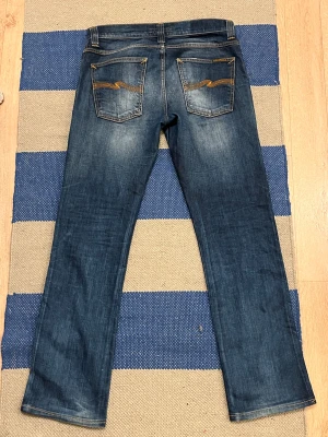 Blå jeans från Nudie Jeans Co, W31 L34 - Snygga blå jeans från Nudie Jeans Co med klassisk femficksdesign och coola orangea sömmar på bakfickorna. Jeansen har en rak passform och är tillverkade i slitstarkt denim. Perfekt för dig som gillar en tidlös och avslappnad stil. Riktigt fin design och bra skick! Storlek: 31/34, hör av dig gällande frågor.