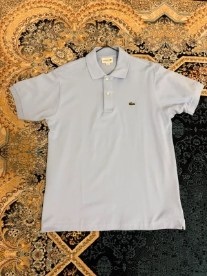 Ljusblå pikétröja från Lacoste - Säljer en klassisk ljusblå pikétröja från Lacoste med broderad krokodil på bröstet. Tröjan har korta ärmar, krage och två knappar framtill. Perfekt för en clean och stilren look.