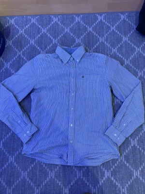 Randig skjorta från Tommy Hilfiger - Säljer en klassisk blå- och vitrandig skjorta från Tommy Hilfiger. Skjortan har lång ärm, knappar framtill och en liten broderad logga på bröstet. Tillverkad i mjuk bomull och har en snygg krage. Perfekt för en clean och stilren look.