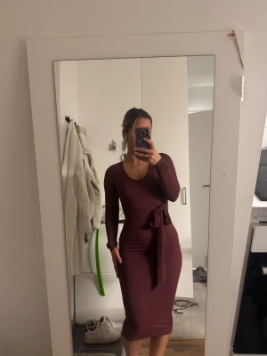 Vinröd ribbad klänning med cut out - Vinröd, ribbad klänning i figurnära modell med långa ärmar och snygga cut out-detaljer på axlarna. Klänningen har en knytning i midjan som markerar siluetten och finns i både midi- och kortare längd. Perfekt för dig som gillar stilrena men trendiga plagg.