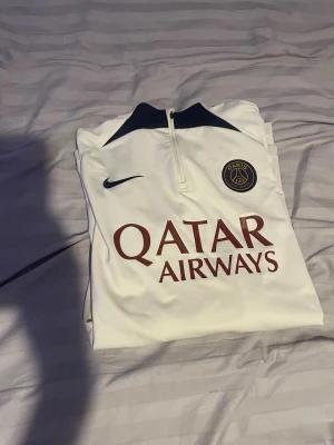 Vit PSG långärmad fotbollströja Nike - Säljer en vit långärmad PSG fotbollströja från Nike med mörkblå detaljer vid kragen och PSG-emblem på bröstet. Tröjan har dragkedja vid halsen och Qatar Airways-tryck på framsidan. Tillverkad i ett lätt och funktionellt material, perfekt för träning eller match.