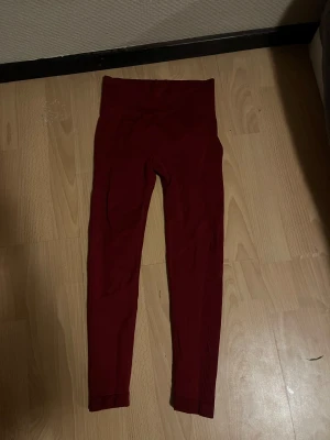Mörkröda scrunch leggings, S - Säljer ett par mörkröda leggings med scrunch-detalj bak och hög midja. Materialet är stretchigt och består av 90% polyamid och 10% elastan, vilket gör dem riktigt sköna och följsamma. Perfekta för gymmet eller chill hemma.