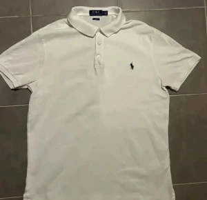 Vit pikétröja från Polo Ralph Lauren - Vit klassisk pikétröja från Polo Ralph Lauren med korta ärmar och krage. Tröjan har tre knappar framtill och den ikoniska blå broderade loggan på bröstet. Tillverkad i mjuk bomull som är skön mot huden och passar perfekt till en clean och fräsch stil.