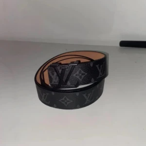 Svart skärp i skinn - Säljer ett svart bälte i skinn med klassiskt monogrammönster i svart metall. Insidan är beige och bältet har tydliga sömmar längs kanterna. Perfekt accessoar för att lyfta din outfit.