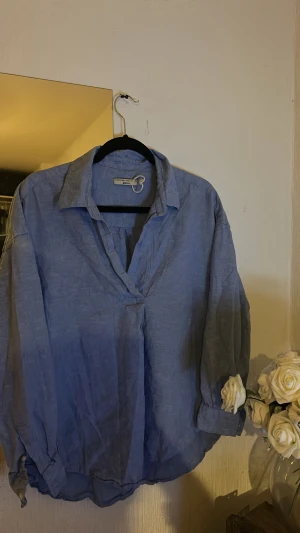 Blå linneskjorta  - Blå skjorta i luftig linnemix med klassisk krage och v-ringad hals. Skjortan har lång ärm med uppvikta muddar och en avslappnad, loose passform. Perfekt för dig som gillar chill och stilren vibe.