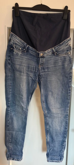 Blå skinny mammajeans från H&M - Säljer ett par blå mammajeans från H&M i storlek L. Modellen är skinny med hög ribbad midja och har en mjuk stretchpanel över magen. Klassisk femficksdesign, dragkedja och knapp framtill. Jeansen är i tvättad blå nyans med lätt slitningar.