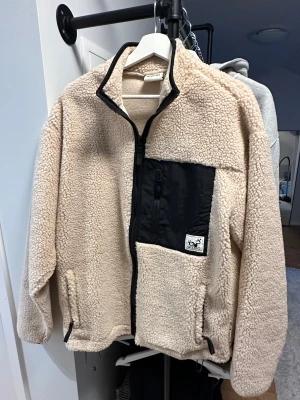 Beige teddyjacka från Gizmo, stl S - Mysig beige teddyjacka från Gizmo med svart bröstficka och dragkedja framtill. Jackan har hög krage, svarta detaljer och en patch med Gizmo-logga på bröstet. Perfekt för kyliga dagar och riktigt skön att bära. Helt ny!