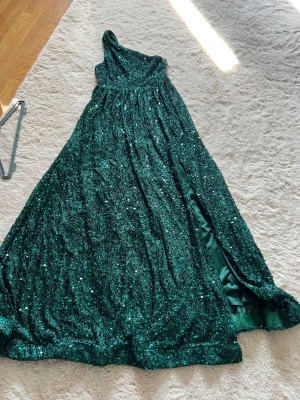 Grön glittrig långklänning oneshoulder - Magisk grön långklänning från KappAhl med glittriga paljetter över hela klänningen. Snygg oneshoulder-modell och en slits på sidan som ger extra edge. Perfekt för dig som vill sticka ut och glänsa. Fodrad insida för skön känsla hela kvällen.