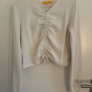 Vit ribbad topp från Gina Young - Säljer en stilren vit ribbad tröja från Gina Young. Toppen har långa ärmar och en fin dragsko framtill som ger en unik look. Perfekt för att lyfta din vardagsstil! Säljer för att den inte kommer till användning💕
